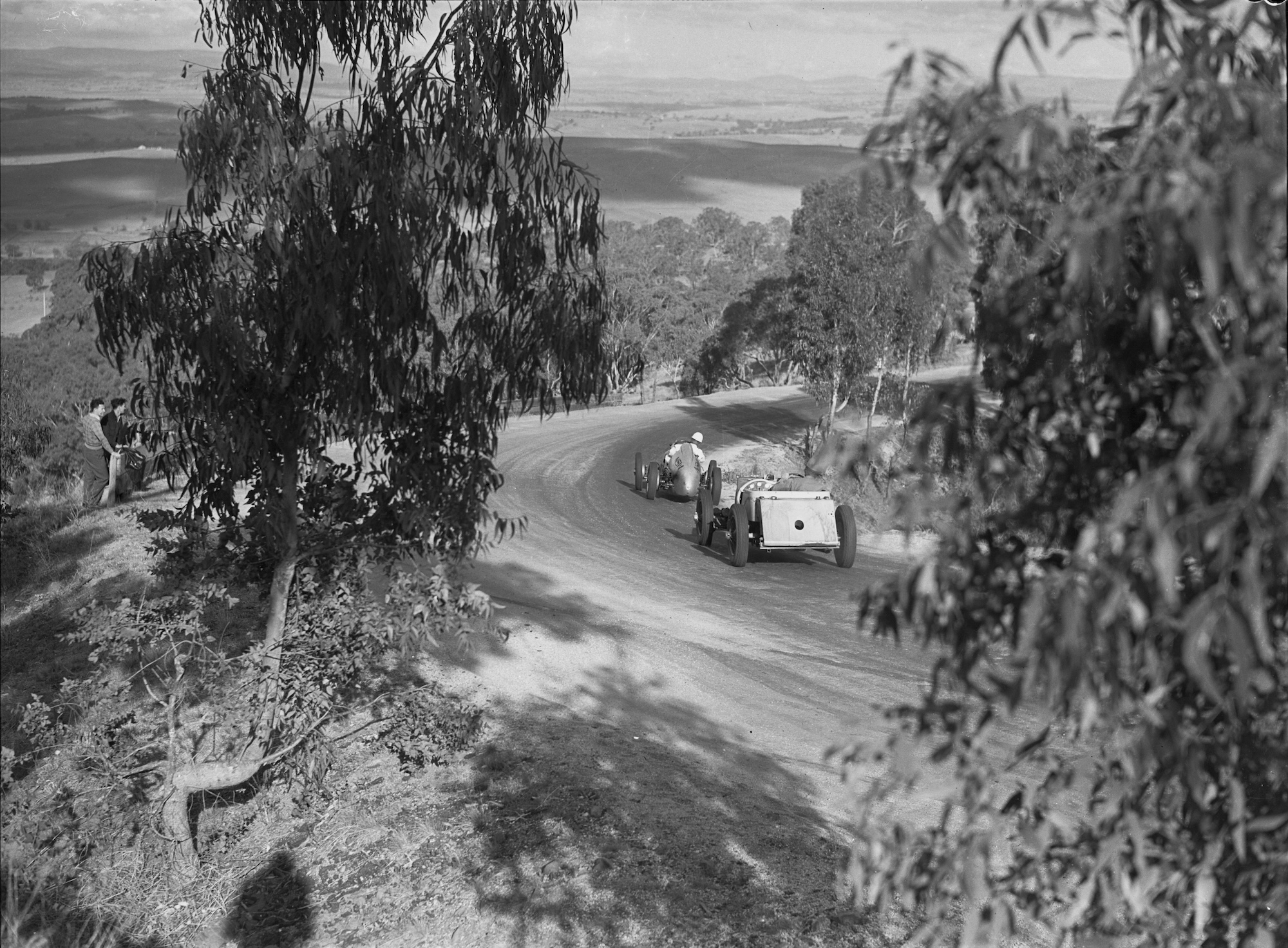 Black-and-white photograph of two open-wheel racing cars negotiating a sweeping bend near the summit of Mount Panorama during the 1946 Australian Grand Prix. The cars race along the narrow, unsealed mountain road, surrounded by eucalyptus trees, with a handful of spectators watching from the roadside and panoramic views of the Bathurst countryside in the distance.